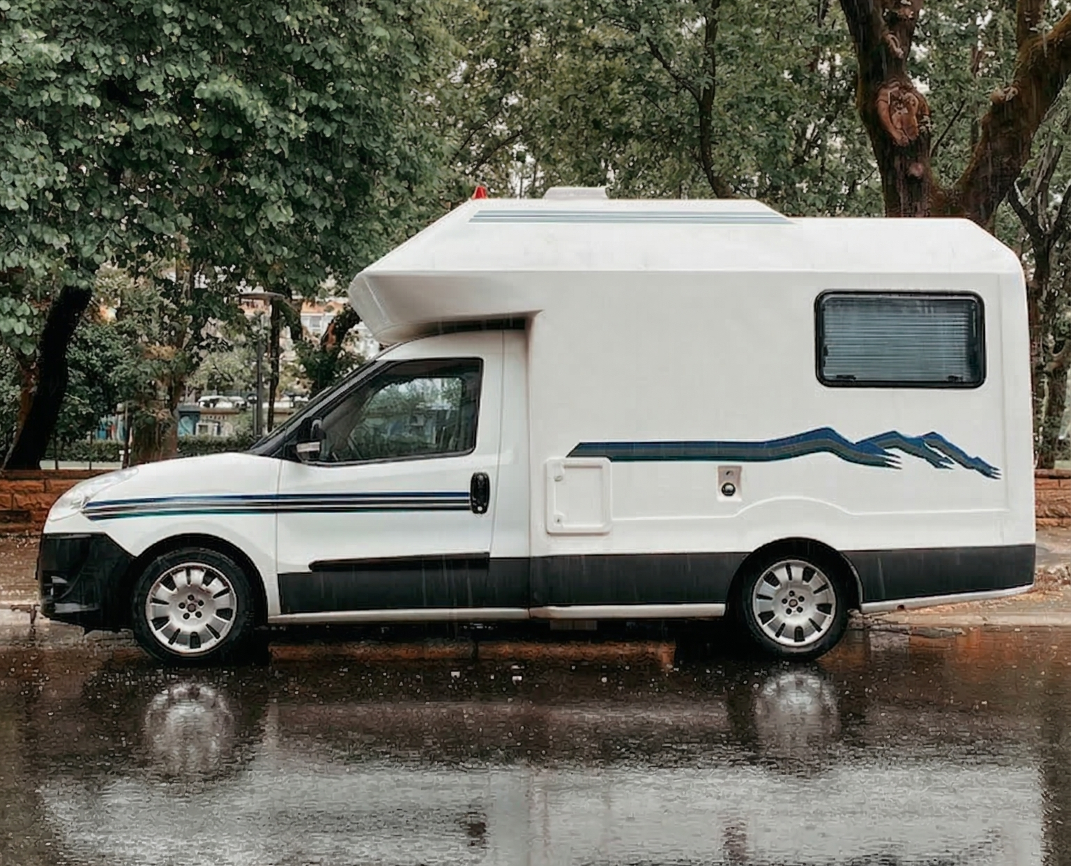 White campervan with mountain decal parked on a rainy street with trees in the background