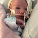 Newborn baby wrapped in pink blanket with a feeding tube resting on hospital bed