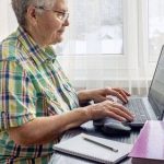 Elderly woman using a laptop at a desk near a window, with a notebook and folder, wearing glasses and casual clothing